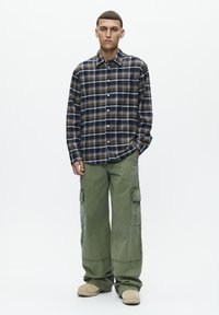 Skjorte i tern i blå, brun og creme; knap-ned design. Cargo-bukser i olivengrøn med sidelommer. Beige slip-on sko.