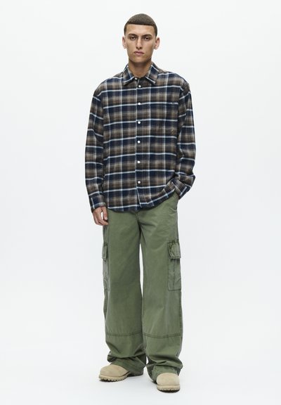 Chemise à carreaux bleue, marron et crème ; design à boutons. Pantalons cargo vert olive avec poches latérales. Chaussures beige à enfiler.