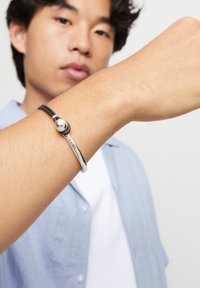 Joven con una pulsera plateada y negra que presenta una cuenta redonda y una barra de metal curvada, levantando el brazo frente a su cara.