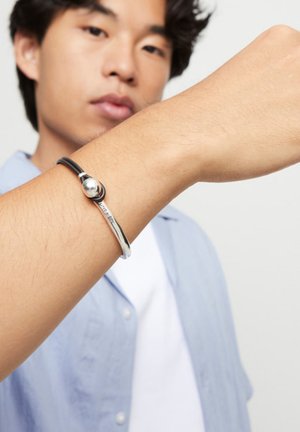 Joven con una pulsera plateada y negra que presenta una cuenta redonda y una barra de metal curvada, levantando el brazo frente a su cara.