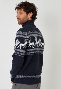 Marineblauer gestrickter Pullover mit hohem Kragen, der weiße Rentiere und geometrische Muster über die Brust zeigt. Weiche Textur, gerippte Bündchen.