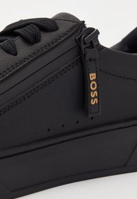 Baskets en cuir noir avec des lacets texturés et une tirette de fermeture éclair latérale portant l'inscription "BOSS" en lettres couleur beige.