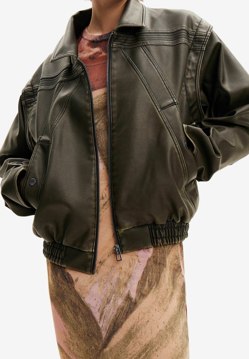 Olive green bomber jacket in smooth fabric, featuring zip closure, ribbed hem, and detailed stitching on shoulders; worn over a printed dress.