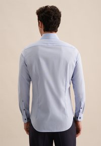 Camicia azzurra chiaro con maniche lunghe, orlo curvo e colletto arrotondato. Presenta bottoni scuri e una texture liscia e tessuta.