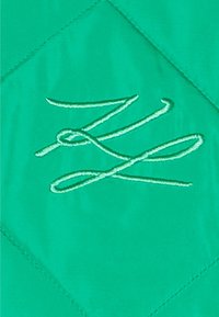 Ljust grönt kviltat tyg med ett diamantmönster, med en ljusgrön broderad logotyp i kursiv i mitten.