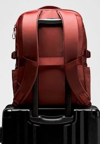 Piros hátizsák rögzítve a fekete gurulós bőrönd fogantyújára, látható párnázott pántokkal és állítható oldalszíjakkal.