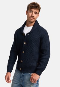 Cárdigan de punto azul marino con cuello de chal, botones de madera, puños y dobladillo acanalados; combinado con jeans desgastados de color azul claro.