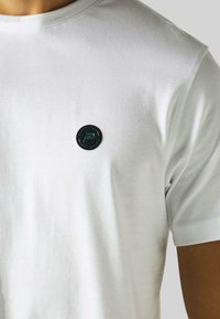 White cotton t-shirt featuring a circular, black emblem with a teal accent on the left chest area. Smooth texture and crew neckline.
