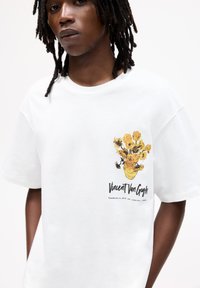 Weißes Baumwoll-T-Shirt mit einem lebhaften Sonnenblumenmotiv und schwarzem Text, kurzen Ärmeln und einem lockeren Schnitt. Glatte Textur.
