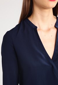 Blouse bleu marine avec une texture douce et lisse, un col en V et des manches longues. Le tissu reflète subtilement la lumière, rehaussant ainsi le design.