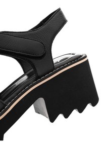 Sandale en cuir noir avec semelle compensée épaisse et semelle crantée, avec une bride Velcro à la cheville et une bordure beige le long du bord de la semelle.