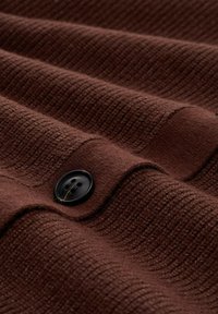 Bruine gestructureerde stof met verticale ribbels, voorzien van een zwart knoopje met twee gaten. Het materiaal lijkt zacht en heeft een warme, uitnodigende toon.