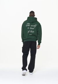 Vihreä huppari, jossa valkoinen teksti "Maailma on väsynyt vihaan", yhdistetty mustiin housuihin ja valkoisiin lenkkareihin, nähtynä takaa.