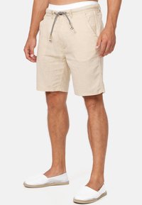 Beige drawstring shorts för män, bärs med vita slip-on skor, visas från midja till fötter mot en vit bakgrund.