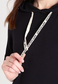 Czarna bluza z kapturem z regulowanym sznurkiem, w kolorze kremowym z nadrukowanym tekstem. Materiał wydaje się miękki i ma luźny krój.