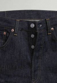 Jeans de mezclilla oscura con cintura alta, diseño de cinco bolsillos y cierre de botón con cinco botones metálicos. Se aprecian acentos de costura de contraste.
