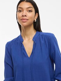 Plava košulja dugih rukava s V-izrezom i suptilnim nabranim detaljima. Izrađena od mekog materijala, ima ležeran kroj i čisti dizajn.