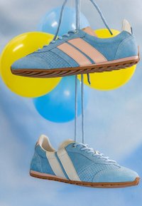 Baskets en daim bleu avec semelles en gomme et bandes latérales contrastées suspendues par les lacets sur un fond de ballons jaunes et bleus.