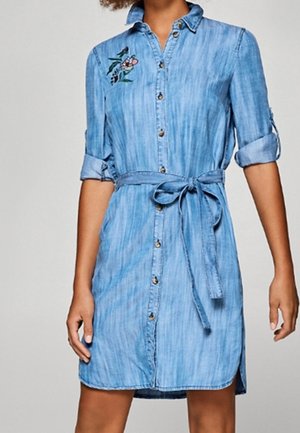 Hellblaues Denim-Hemdkleid mit hochgekrempelten Ärmeln, Knöpfen vorne, Taillengürtel und kleiner Blumenstickerei auf der oberen linken Seite.