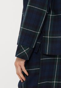 Μανίκι σακακιού σε ταρτάν σε σκούρο πράσινο και ναυτικό με λευκές λεπτομέρειες. Το ύφασμα φαίνεται υφή, με κομμένη εφαρμογή και λεπτομέρεια στο μανίκι.