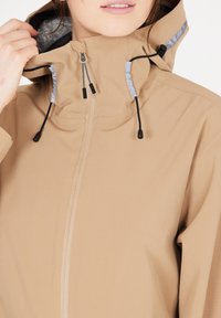 Beige vindjakke med lynlås, justerbar snoretræk-hood med grå fòr og glat, let stofstruktur.
