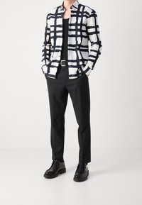 Chemise blanche à motif quadrillé noir, col boutonné, associée à un pantalon noir et des chaussures. Chaussures plates légèrement brillantes, design minimaliste.