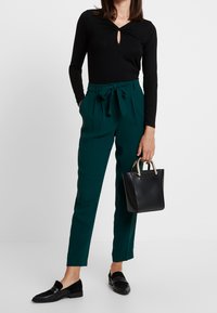 Černý top s dlouhými rukávy a detaily ve tvaru klíče, zkombinovaný s vysokými zelenými kalhotami s uvazovacími pásky. Modelka nese malou černou kabelku.