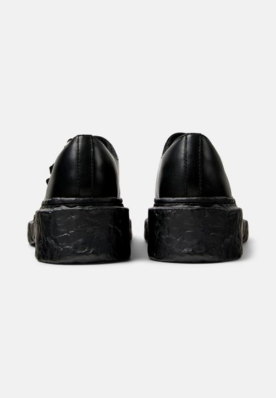 Zapatos de cuero negros con una suela de goma texturizada y gruesa, mostrando una vista trasera. El diseño incluye costuras mínimas y un acabado suave.