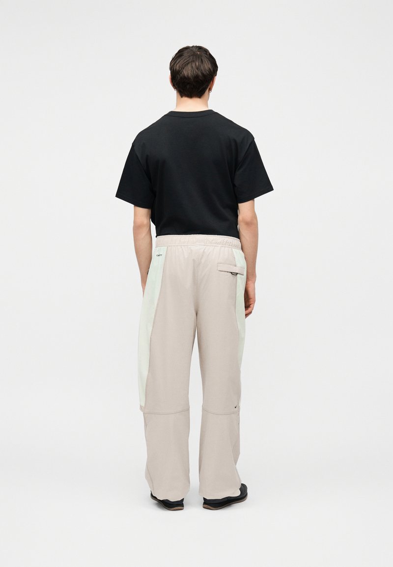 Man staat met de rug naar de camera, draagt een zwart shirt met korte mouwen en losse beige broek met lichte zijpanelen en achterzak, tegen een effen achtergrond.