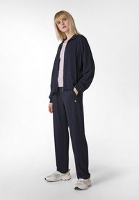 Giacca di tuta blu navy con colletto alto e polsini elastici, abbinata a pantaloni larghi coordinati. Maglietta rosa chiaro sotto e sneakers.