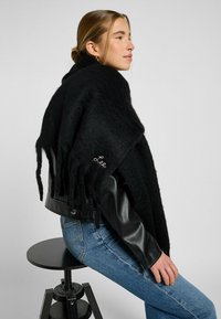 Châle à franges noires en laine, avec une finition texturée et un détail brodé "Lee", porté sur une veste en cuir noire et un jean bleu.