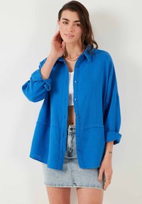 Blå, button-up skjorte i tekstureret stof, lange ærmer rullet op ved manchetterne og to forlommer. Kombineret med en hvid top og en denimnederdel.