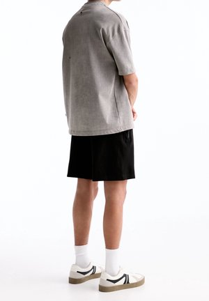 Camiseta gris de manga corta con un ajuste relajado, combinada con pantalones cortos negros. El calzado presenta detalles en blanco y negro sobre una suela clara.