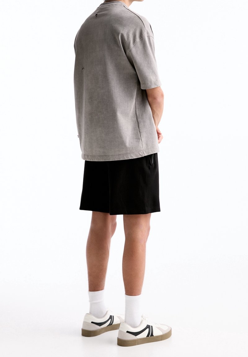 Chemise grise à manches courtes avec une coupe décontractée, associée à un short noir. Les chaussures présentent des accents blancs et noirs sur une semelle claire.