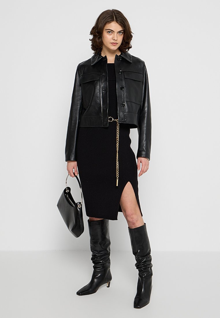 Giubbotto corto in pelle nera con chiusura a bottone, indossato sopra un vestito nero a coste con uno spacco laterale e un accento in catena dorata. Stivali neri al ginocchio e borsa.