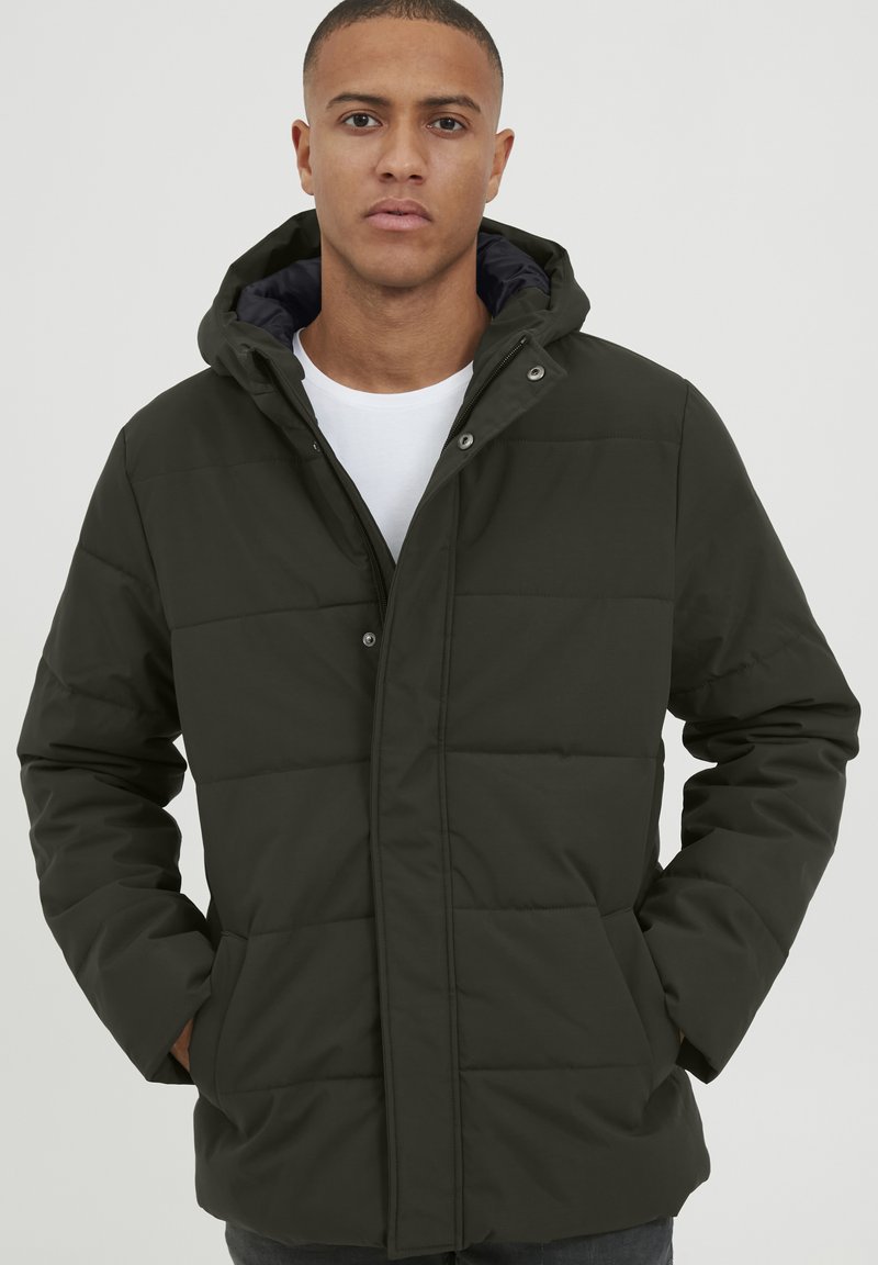 Dunkelgrüne gepolsterte Jacke mit Kapuze, mit Druckknopfverschlüssen, Seitentaschen und einer glatten Textur. Getragen über einem weißen Hemd.