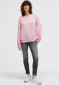 Eine Frau steht vor einem einfarbigen Hintergrund und trägt eine hellrosa Bluse mit langen Ärmeln, graue enge Jeans, die an den Knöcheln hochgerollt sind, und weiße Sneakers.