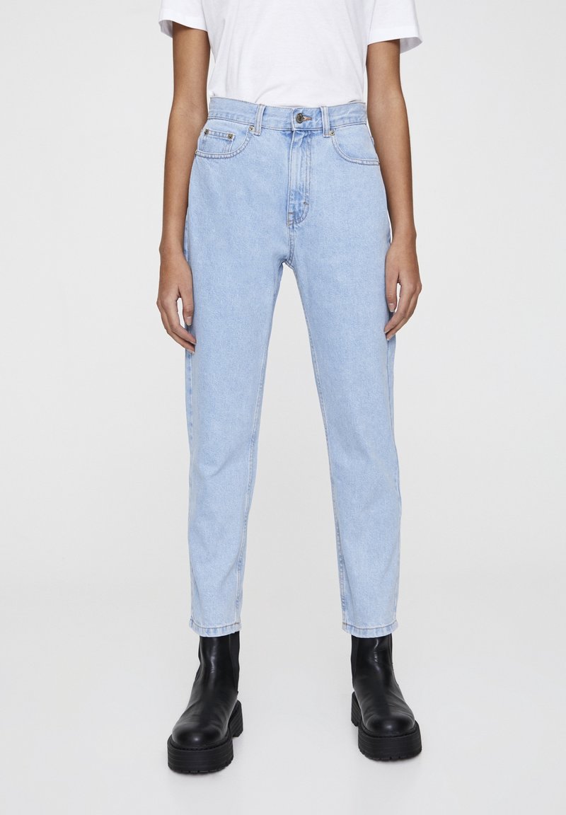 Personne portant un jean droit taille haute bleu clair, une chemise blanche à manches courtes et des bottines noires épaisses, debout devant un fond uni.