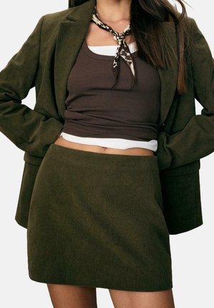 Femme portant un blazer et une jupe en velours côtelé vert olive avec un top court marron sur une chemise blanche et un foulard à motifs autour du cou.
