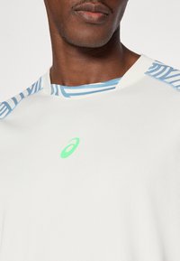 Άσπρο αθλητικό πουκάμισο με λαιμόκοψη σε μπλε μοτίβο και μικρό πράσινο λογότυπο στο στήθος. Υφασμάτινη υφή με ριμπ λεπτομέρειες.