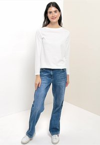 Witte lange mouwen top, relaxte pasvorm, gecombineerd met lichtblauwe rechte jeans en witte sneakers, op een neutrale achtergrond.