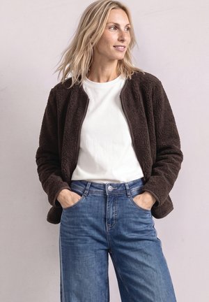 Kobieta ubrana w ciemnobrązową polarową kurtkę na białej koszuli z niebieskimi dżinsami, stoi z rękami w kieszeniach, patrzy w prawo.