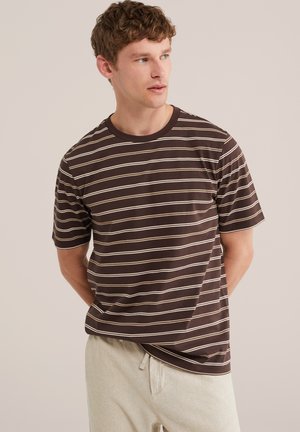 Ung mand iført en brunt og beige striped kortærmet skjorte og lysebeige bukser, stående med hænderne bag ryggen og kigger til siden.