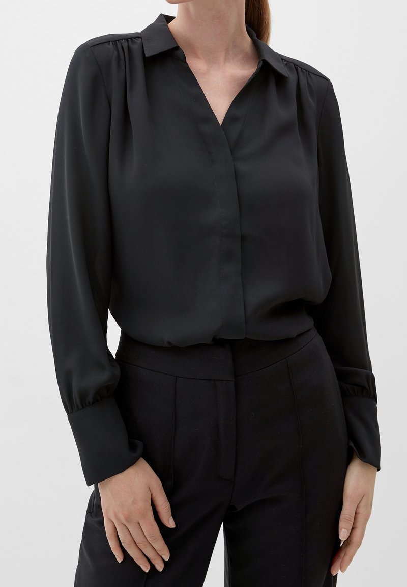Femme portant un chemisier noir à manches longues avec col et un pantalon noir taille haute, mains posées sur les hanches, debout devant un fond uni.