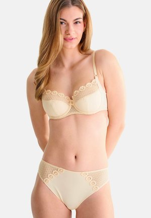 Femme portant un soutien-gorge en dentelle beige et une culotte assortie, debout les mains derrière le dos sur fond blanc.