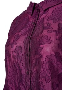 Liila paita, joka on valmistettu teksturoidusta kankaasta ja jossa on kukkakuosi, nappikiinnitys edessä ja kaulus. Sujuva pinta, jossa on hienovarainen kiilto ja monimutkaiset yksityiskohdat.