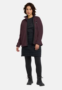 Bordeaux fleecedekenjack met een hoge kraag en een zwarte rits, gedragen over een zwarte puffer rok en zwarte legging. Zwarte wandelschoenen maken de look compleet.