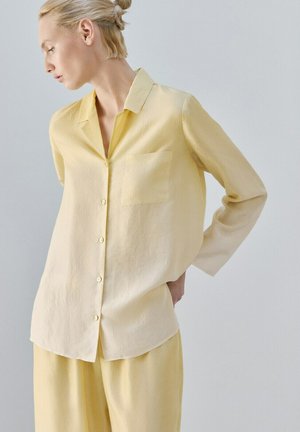 Femeie purtând o cămașă largă galben pal cu nasturi și pantaloni asortați, privindu-se spre stânga, cu părul prins într-un coc.