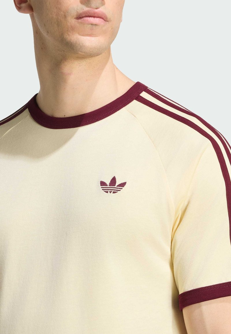 Hombre con camiseta crema con ribete burdeos en el cuello, las mangas, tres rayas en los hombros y el logo de Adidas en el pecho.