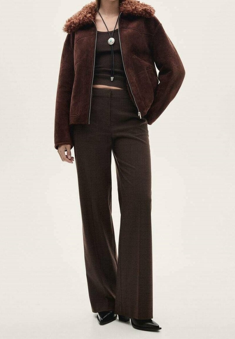Femme portant une veste à col en fourrure marron foncé, un top court côtelé, un pantalon large taille haute, des chaussures noires à bouts pointus et un collier pendentif.